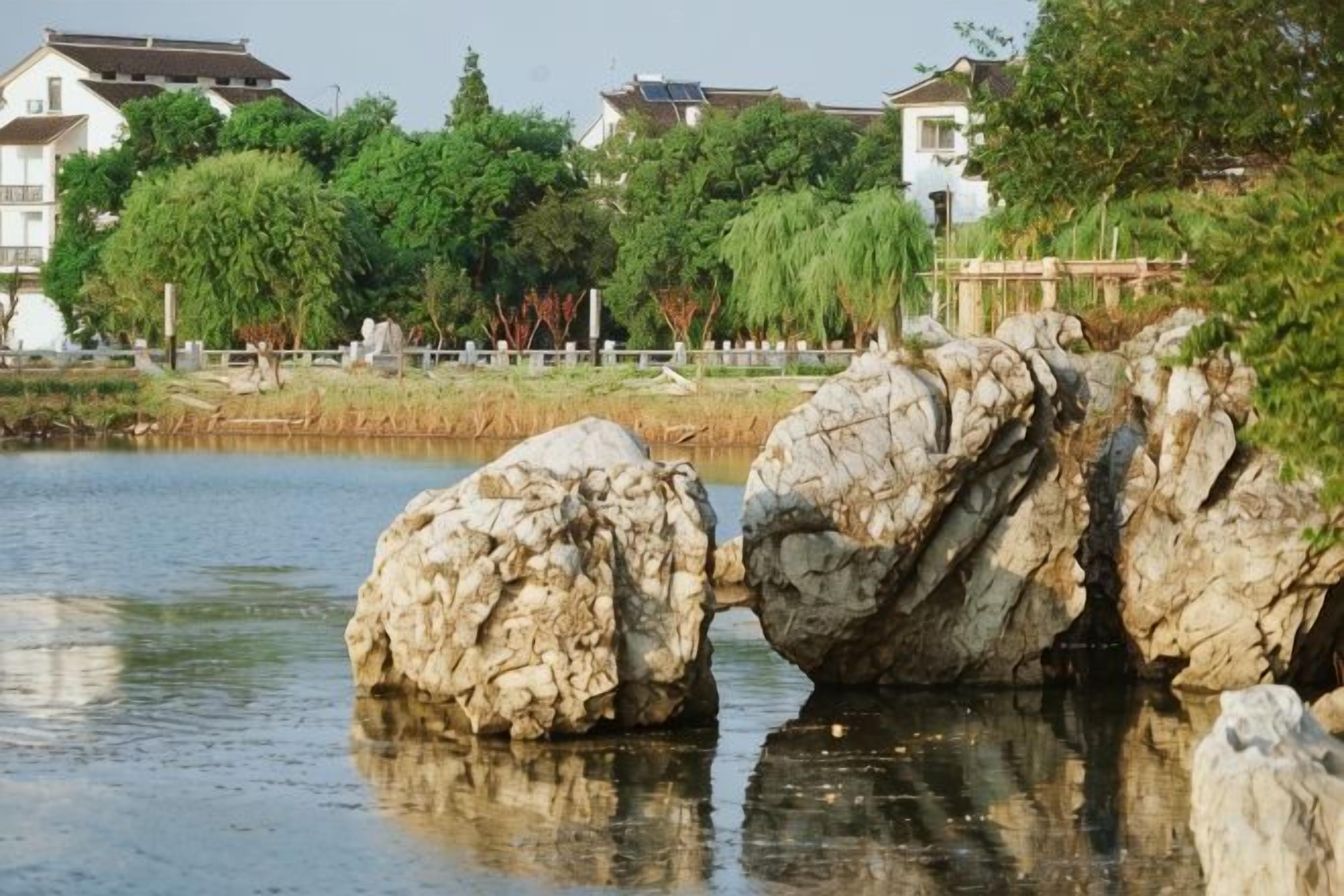 Een rustig waterlichaam met grote rotsen op de voorgrond, een houten omheining en huizen op de achtergrond.