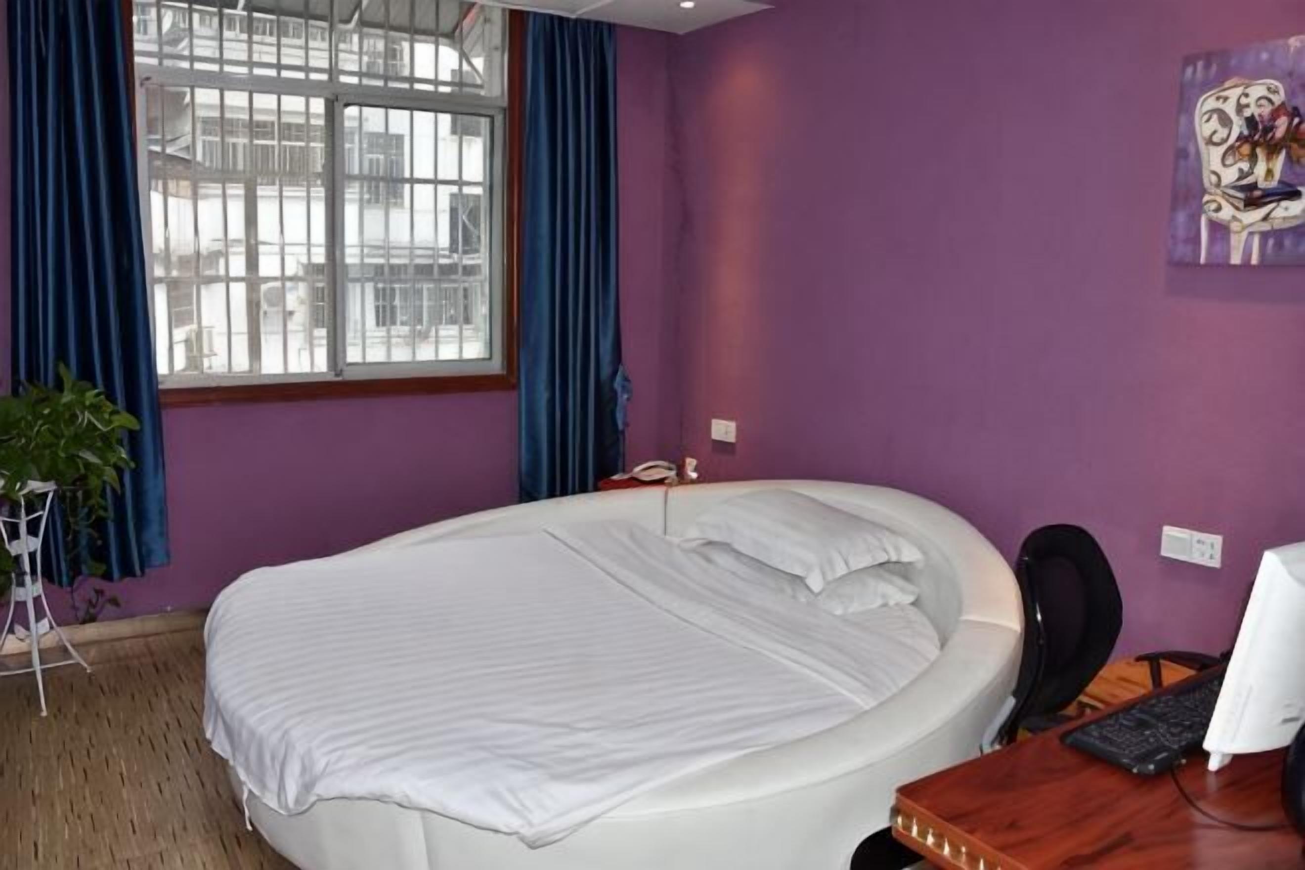 A room with a large bed, a desk with a computer, a chair, a window with curtains, and a plant.