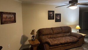 Una sala de estar con un sofá marrón, una pequeña mesita de madera con una lámpara y dos cuadros en la pared.