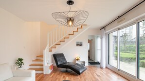 A modern living room with a white sofa, a black lounge chair, a chess table, a wooden staircase, and large windows offering a view of the outdoors. - La Petite Maison Dans la Prairie - Havre de paix (Rennes)