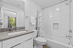 Un baño moderno con lavamanos, inodoro y bañera blancos, cortina de ducha y ventana con cortinas.