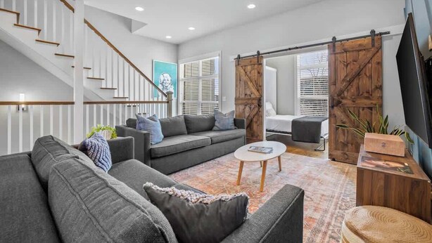 A modern living room with a grey sofa, a coffee table, a staircase with wooden railings, and a sliding barn door. - Rooftop Views! Luxury East Nash 3 BR Townehome (Nashville)