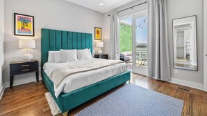 A bedroom with a teal upholstered bed, white bedding, a nightstand with a lamp, a large window with curtains, and a view of greenery outside. - Rooftop Views! Luxury East Nash 3 BR Townehome (Nashville)