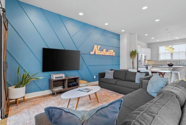 Una sala de estar moderna con una pared azul destacada, un televisor de pantalla plana, un sofá, una mesa de centro y una cocina al fondo.