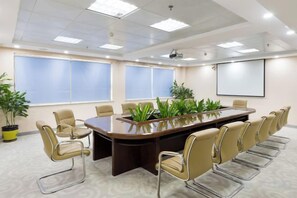 Un salón de conferencias con una larga mesa, sillas, una pantalla de proyector y plantas en macetas.
