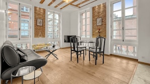 A modern living room with a sofa, dining table, and chairs, large windows, and exposed wooden beams.