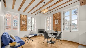 A modern living room with a blue sofa, dining table, and chairs, wooden beams on the ceiling, and large windows with a city view. - Frère Jacques - Appt situé plein centre (Toulouse)