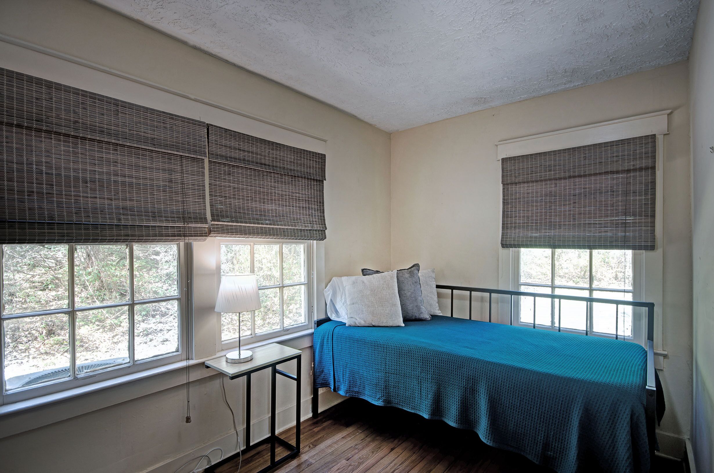 A bedroom with a bed, two windows with blinds, a bedside table with a lamp, and a view of trees outside.