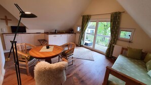 A cozy room with a wooden dining table, chairs, a fluffy chair, a sofa, a ceiling lamp, a TV, a kitchen area, and a window with curtains.