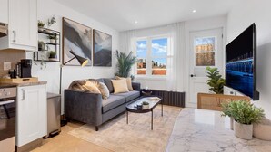 Una sala moderna con un sofá gris, una mesa de centro y un televisor. Hay plantas, un área de cocina con armarios y electrodomésticos, y un ventanal con vistas a la ciudad.