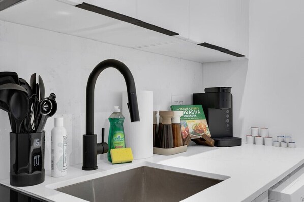 Une cuisine moderne avec un plan de travail blanc, un robinet noir, une machine à café, ainsi que divers ustensiles et appareils de cuisine.