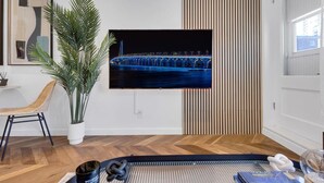 A modern living room with a large flat-screen TV mounted on the wall, a potted plant, a wicker chair, and a wooden floor. - Downtown Montreal Speakeasy 502 | Balcony, Queen Bed, Sofa Bed, Parking & 65` TV (Montreal)