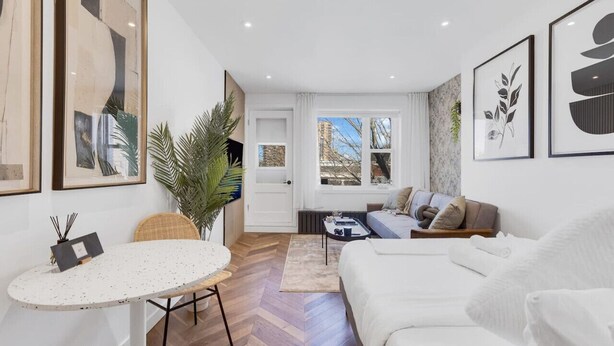 A modern living room with a white sofa, a small round table, and a coffee table. There are framed artworks on the walls and a large window providing a view of the outside. - Downtown Montreal Speakeasy 502 | Balcony, Queen Bed, Sofa Bed, Parking & 65` TV (Montreal)
