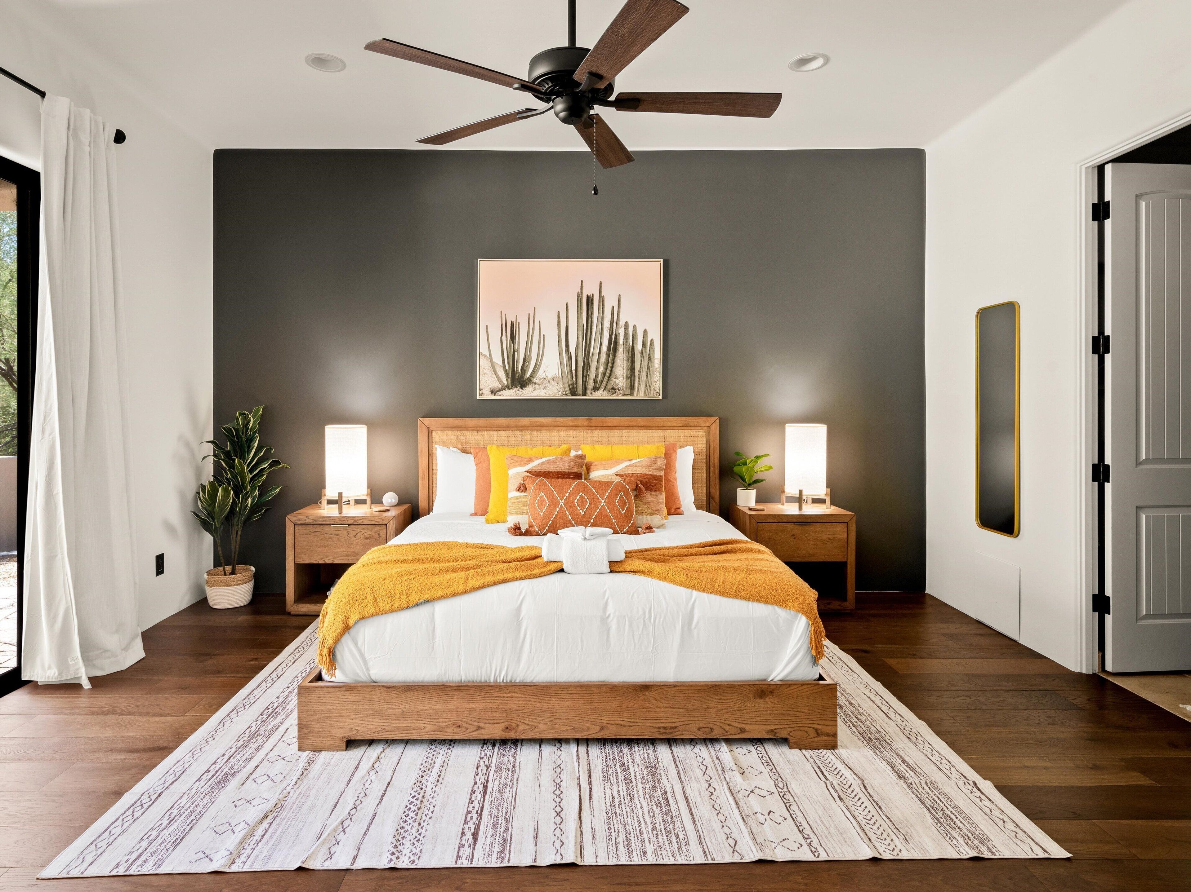 A bedroom with a wooden bed, two bedside tables with lamps, a ceiling fan, a large cactus artwork, and a view of the outdoors.