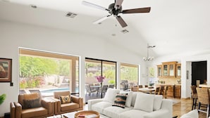 Ein geräumiges Wohnzimmer mit einer großen Couch, einem Couchtisch und einem Essbereich mit Stühlen und Tisch. Es gibt einen Deckenventilator und einen Blick ins Freie durch große Fenster.