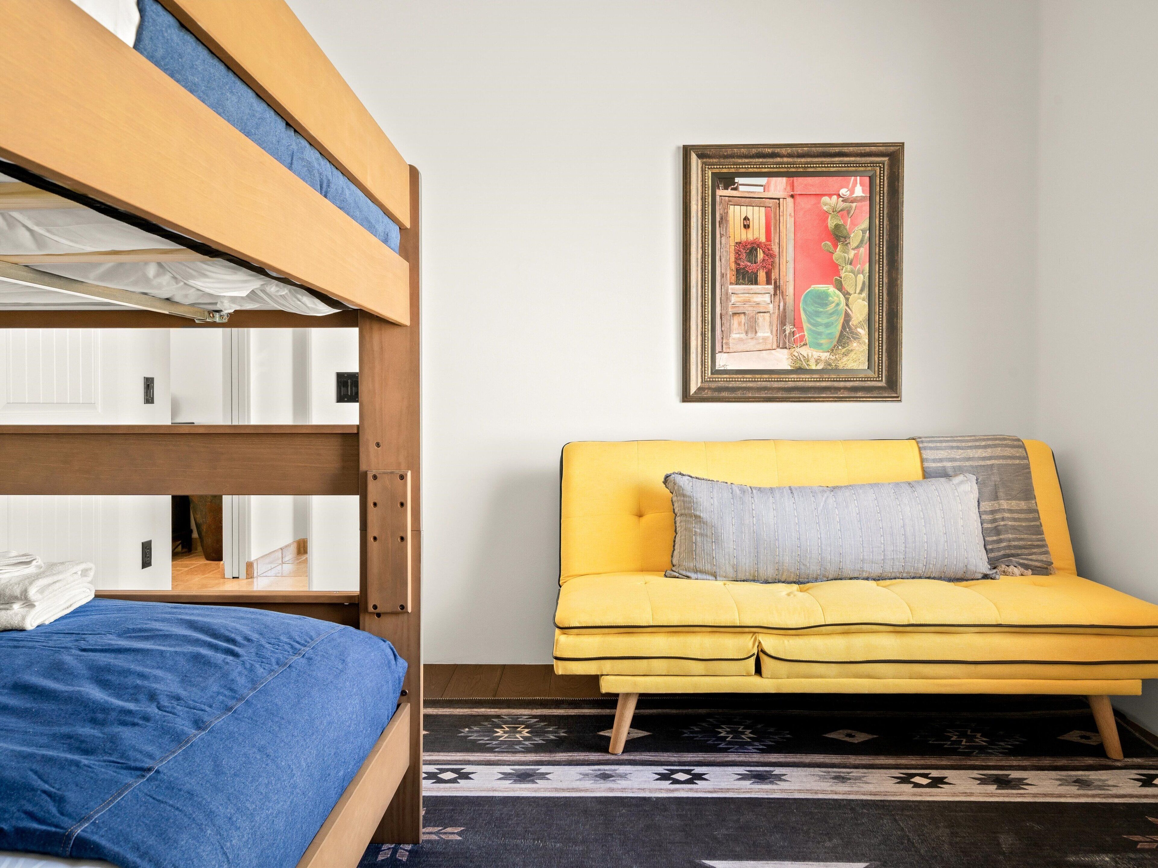 A bunk bed with a blue blanket, a yellow sofa with a gray pillow, and a framed picture on the wall.