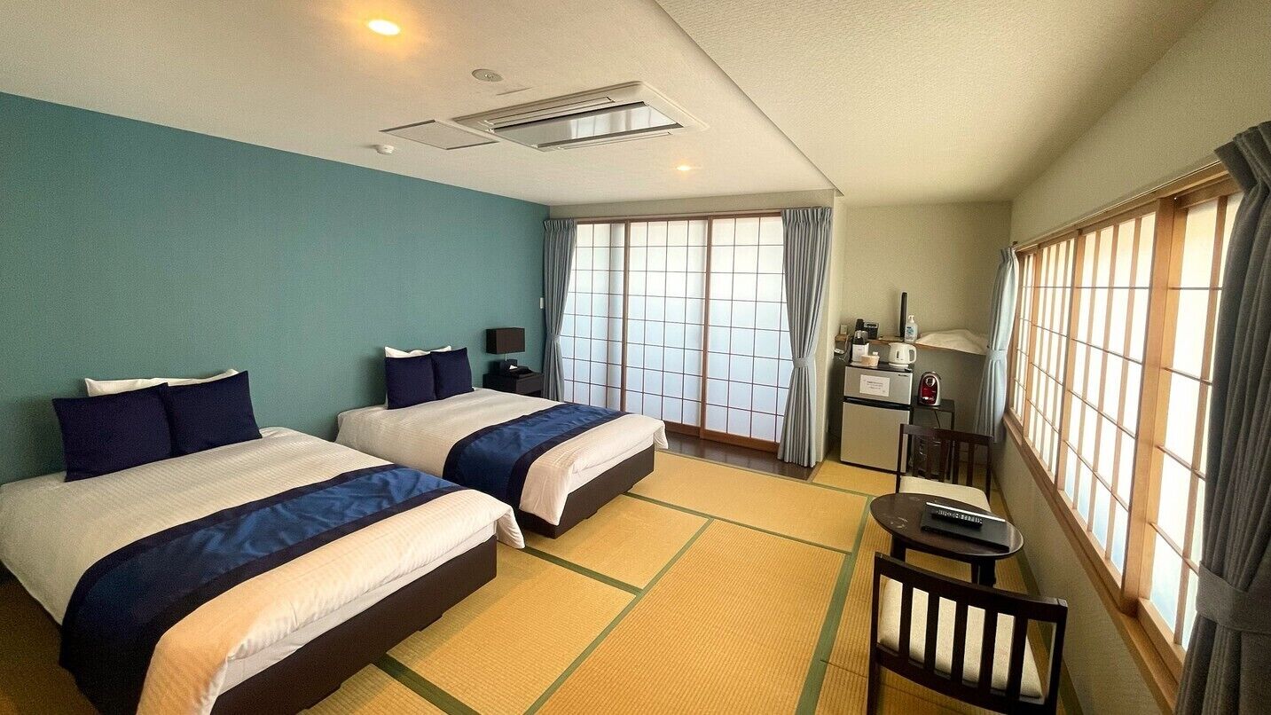 A hotel room with two beds, a small table, a chair, a desk with a lamp, and a window with curtains.
