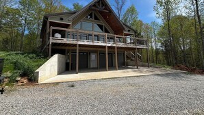 A large, multi-story wooden house with a balcony, set in a forested area with a gravel driveway.