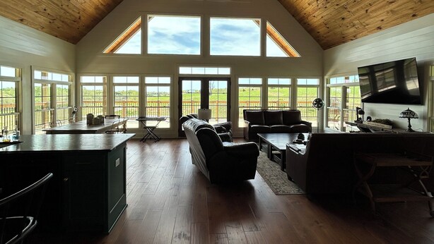 A spacious living area with a high ceiling, wooden beams, and large windows offering views of the outdoors.