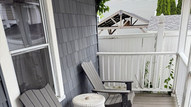 Une véranda couverte avec deux fauteuils Adirondack et une petite table ronde, une clôture blanche et une maison aux murs recouverts de bardage gris.
