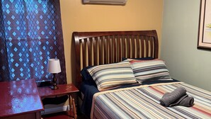 Une chambre avec un lit en bois, une table de chevet avec une lampe, une fenêtre avec des rideaux à motifs et un climatiseur fixé au mur.