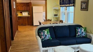Un salon chaleureux avec un canapé et deux fauteuils en osier blancs, un plancher en bois et une cuisine en arrière-plan.