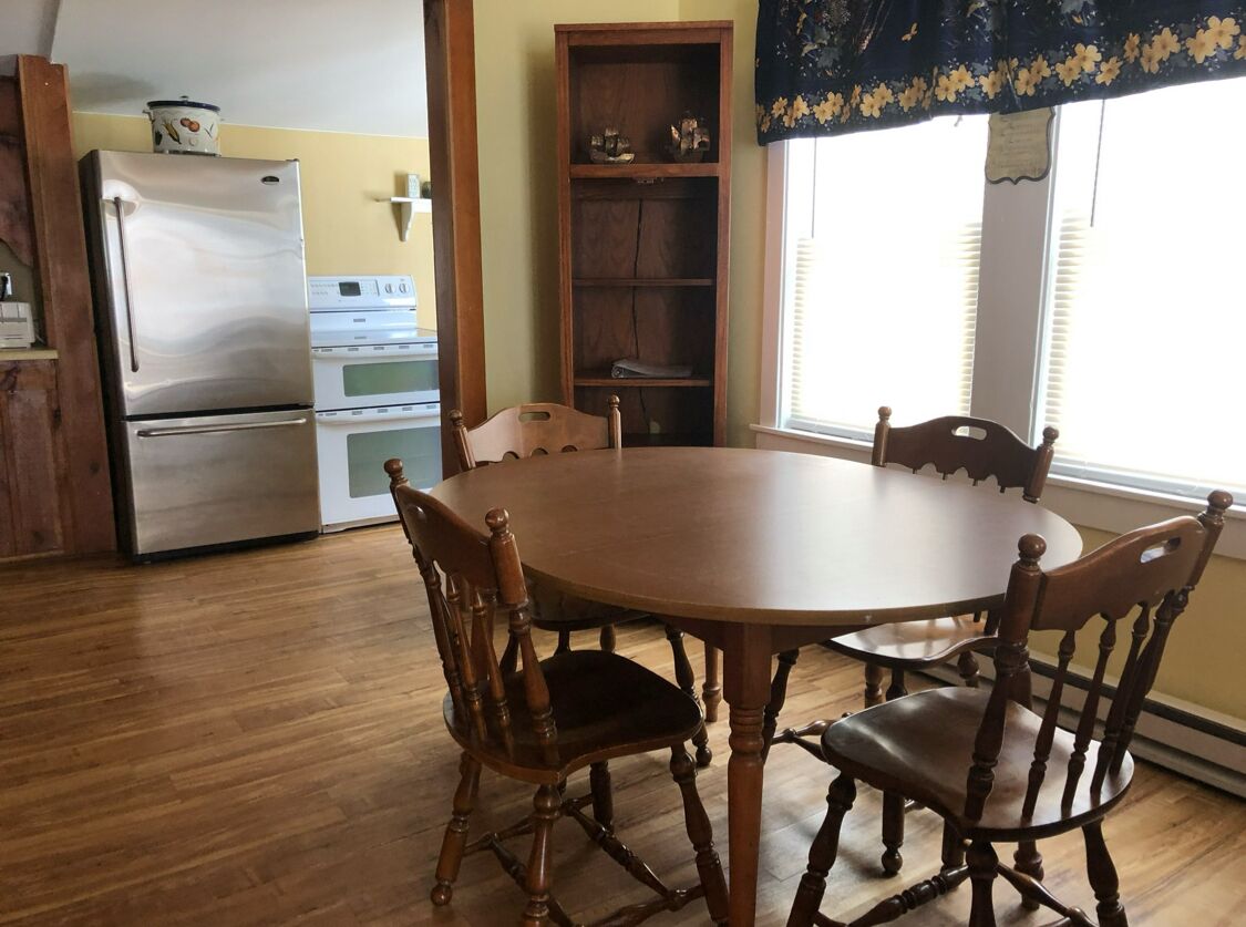 A dining area with a round wooden table and six chairs, a kitchen with a refrigerator and stove, and a window with a floral curtain.