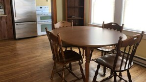 Un coin repas avec une table ronde en bois et six chaises, une cuisine équipée d’un réfrigérateur et d’une cuisinière, et une fenêtre agrémentée d’un rideau à motifs floraux.