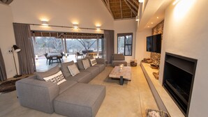 A spacious living area with a sectional sofa, a flat-screen TV, a fireplace, and a large window overlooking a deck. - Villa Appelblaar in Hoedspruit (Hoedspruit)