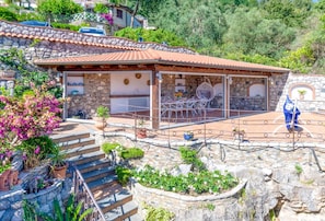 Un bĂątiment en pierre avec un toit en tuiles, un patio couvert avec des meubles dâextĂ©rieur et un escalier en pierre menant Ă lâentrĂ©e.