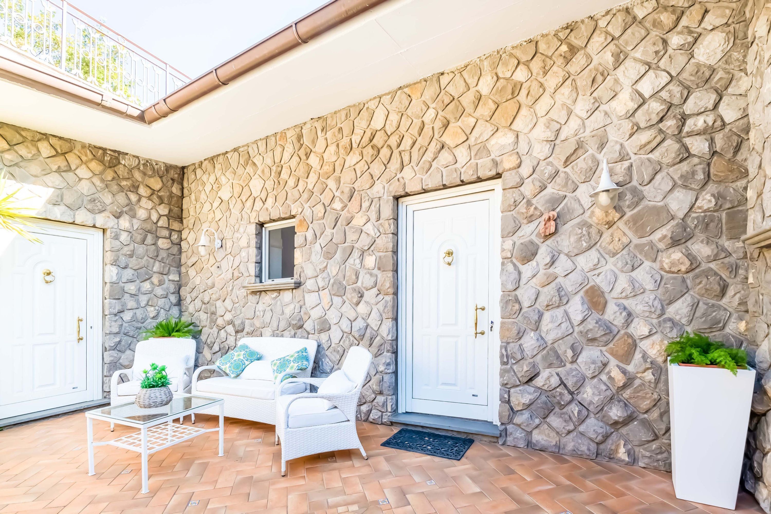 A stone-walled exterior with a white door, a glass table with a plant, and a white chair.
