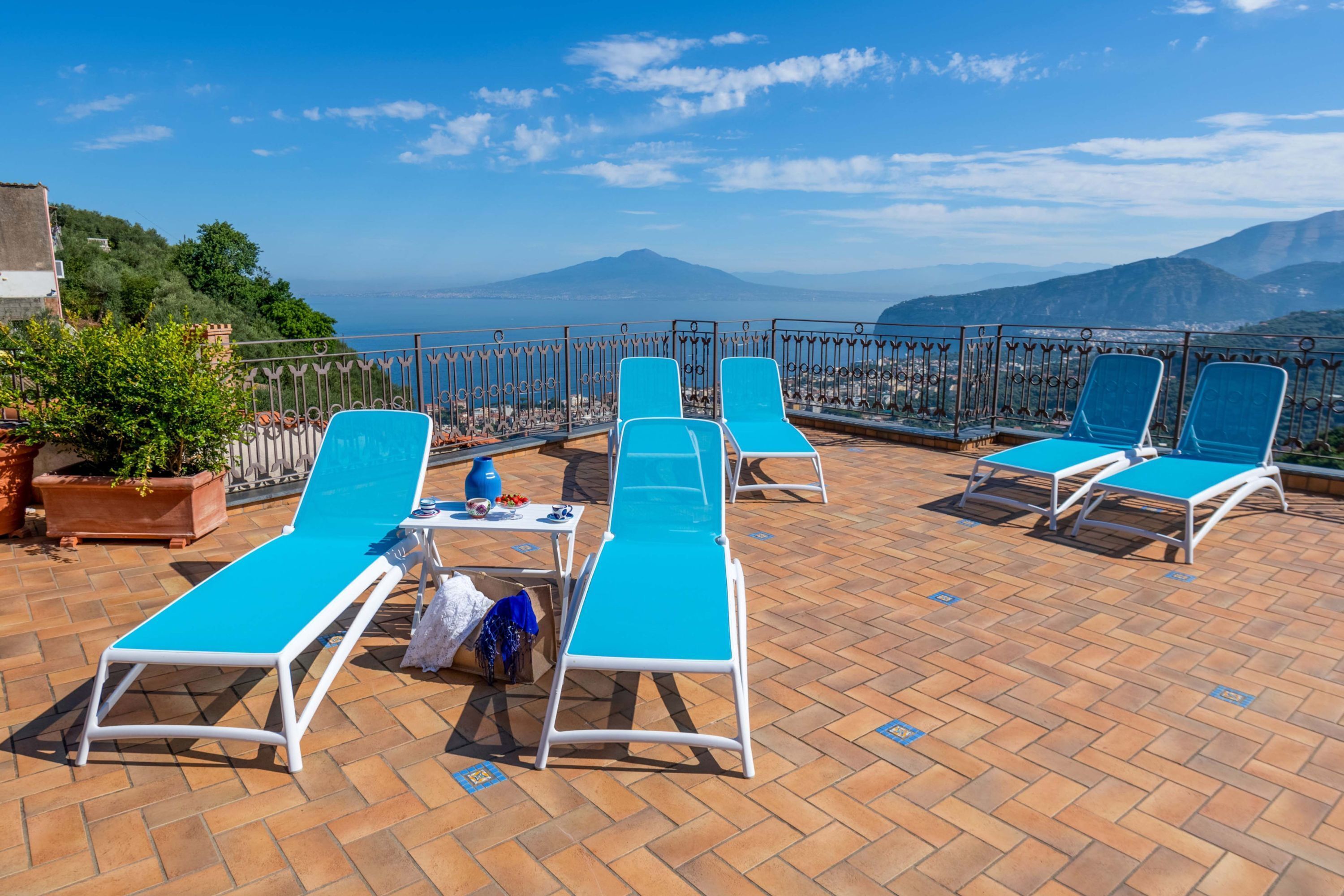 Une terrasse avec des fauteuils bleus, une table ornée d’un vase et d’une bouteille, et une vue sur la mer et les montagnes.