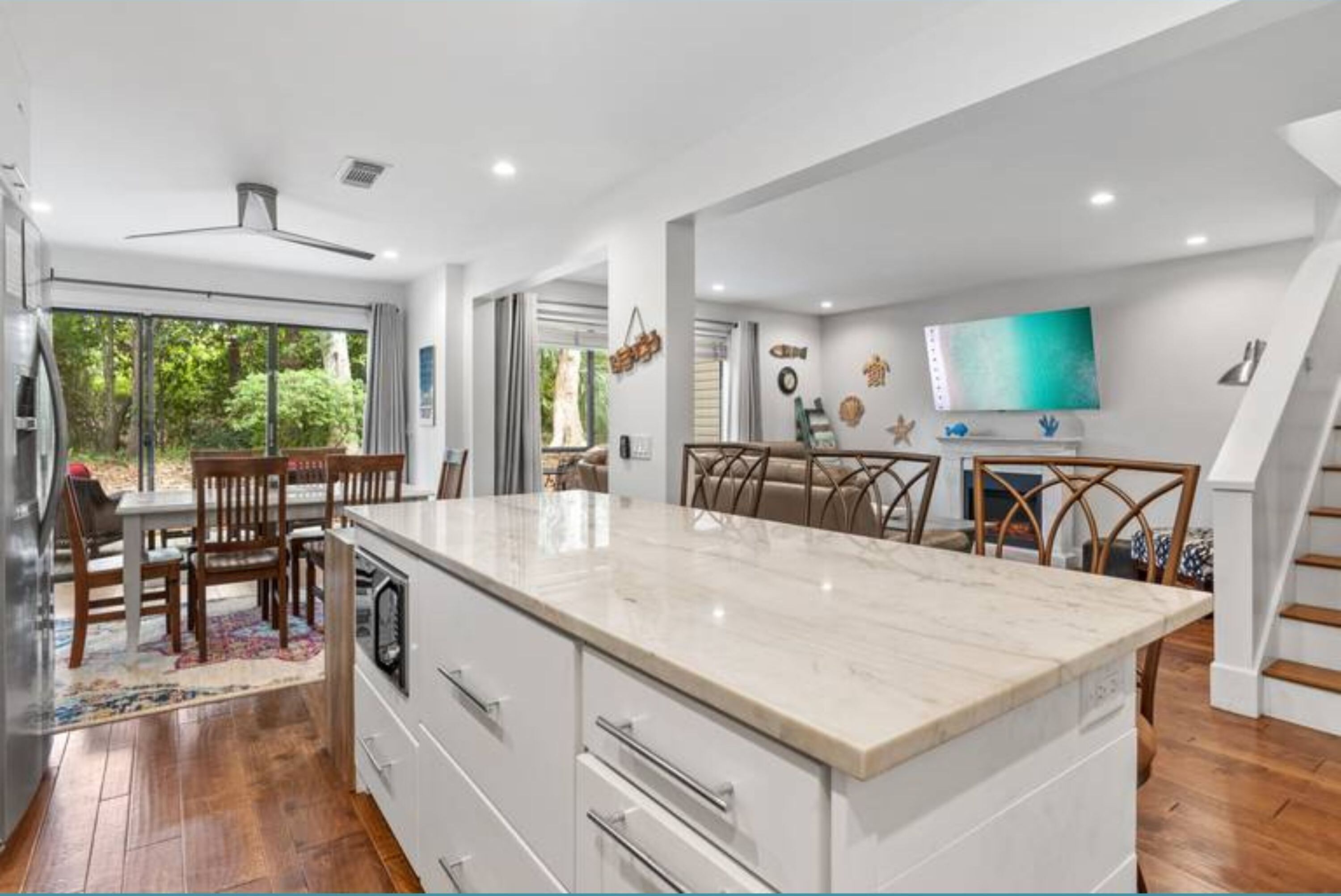 A modern kitchen with a large island, wooden dining table, and chairs. There is a TV mounted on the wall and a staircase leading to another level.