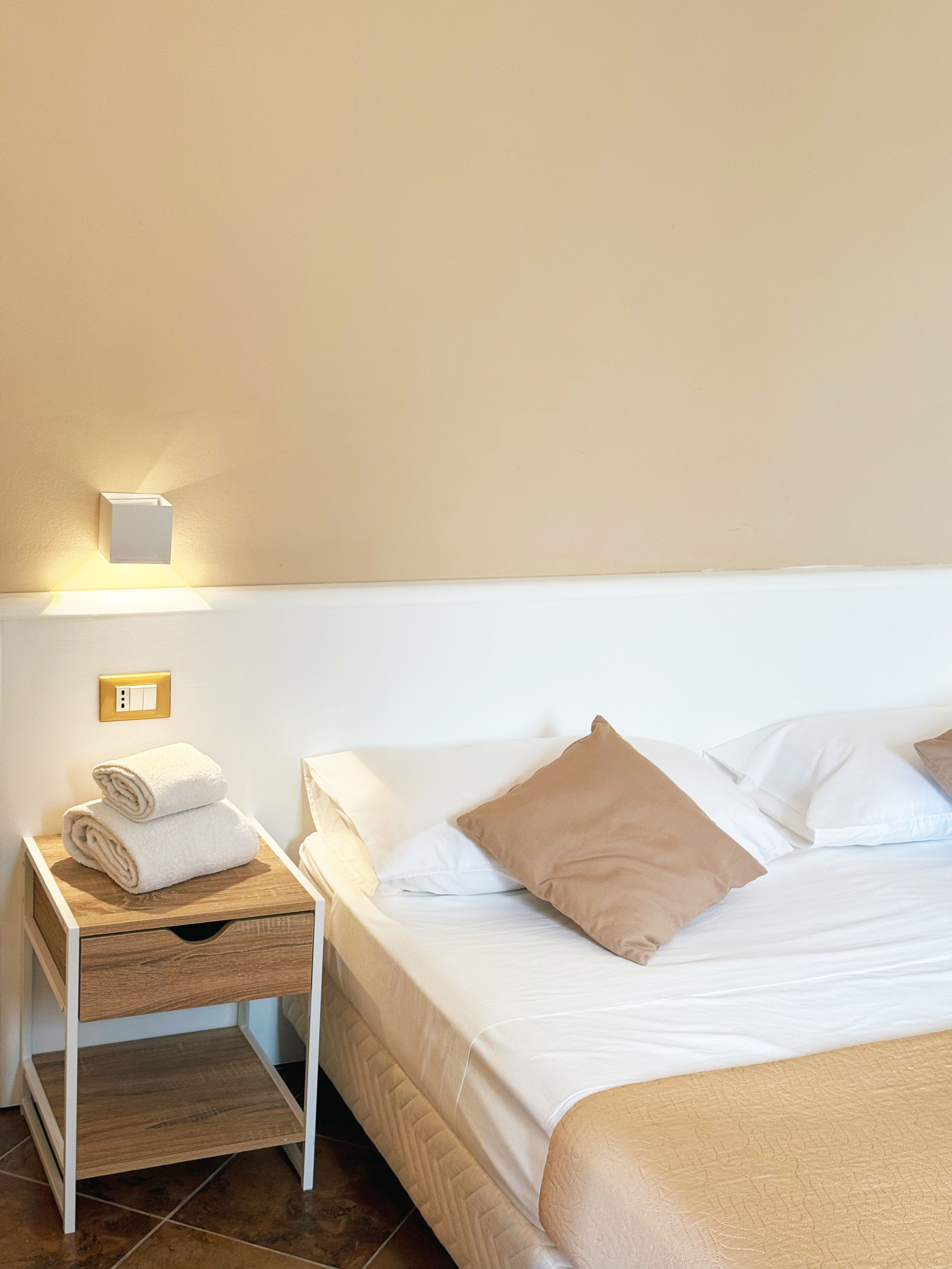 Una cama bien hecha con una manta color crema, una mesita de noche de madera con una toalla doblada, y una lámpara fijada a la pared.