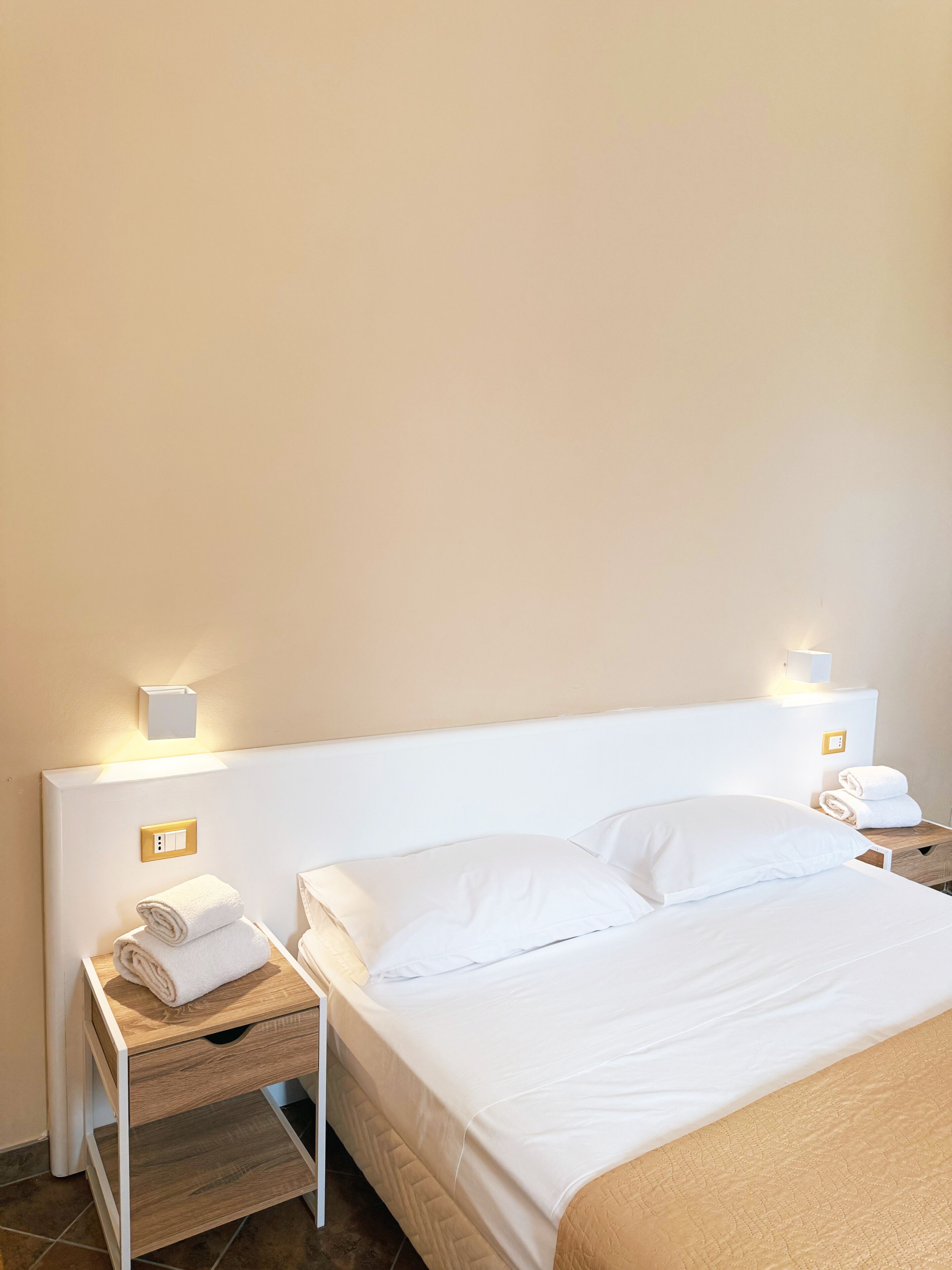 A hotel room with a neatly made bed, two bedside tables with lamps, and a small wooden side table with folded towels.