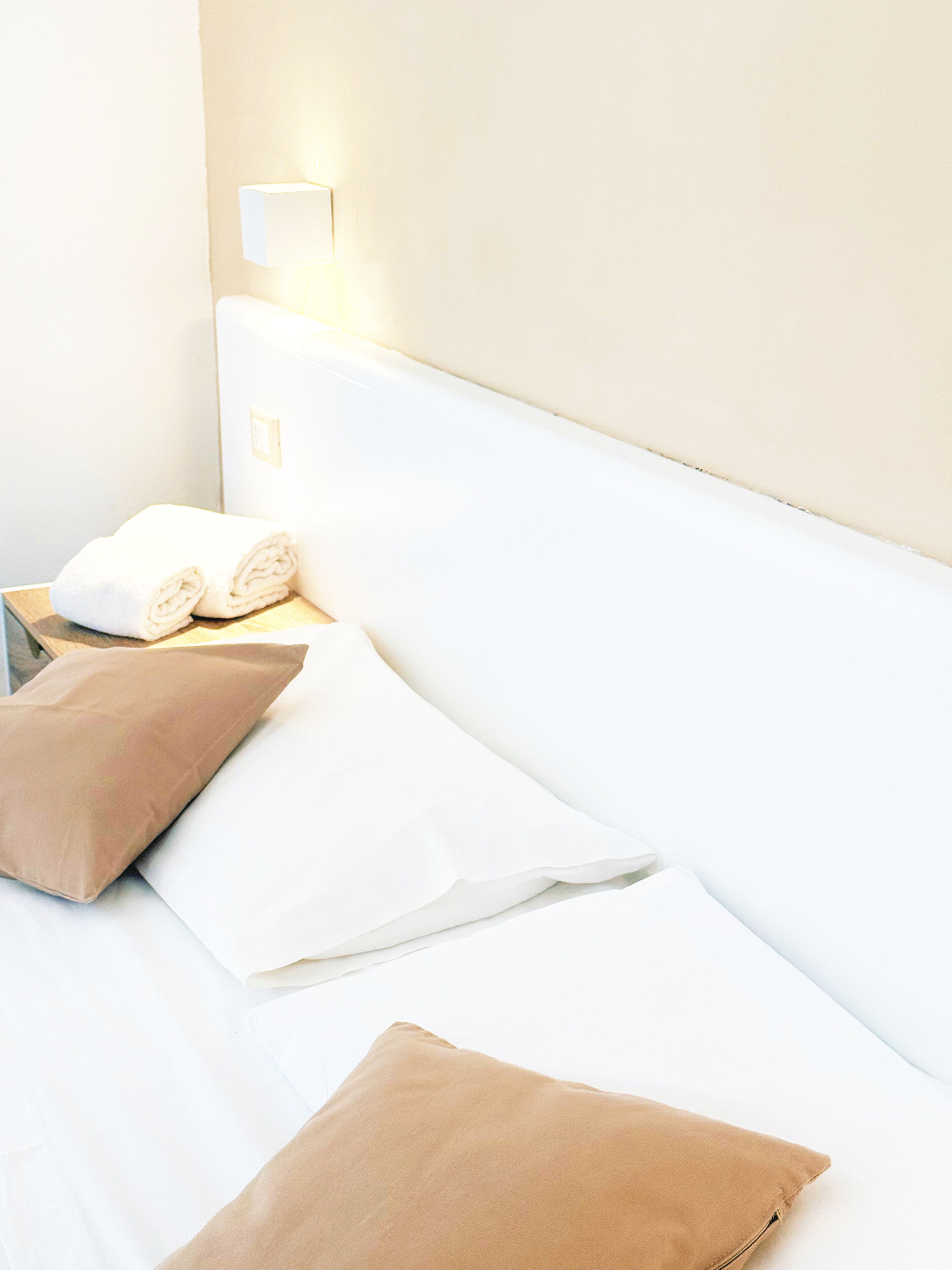 Una cama bien hecha con sábanas blancas y dos almohadas color beige, una lámpara de noche y una mesita de madera con una toalla blanca enrollada.