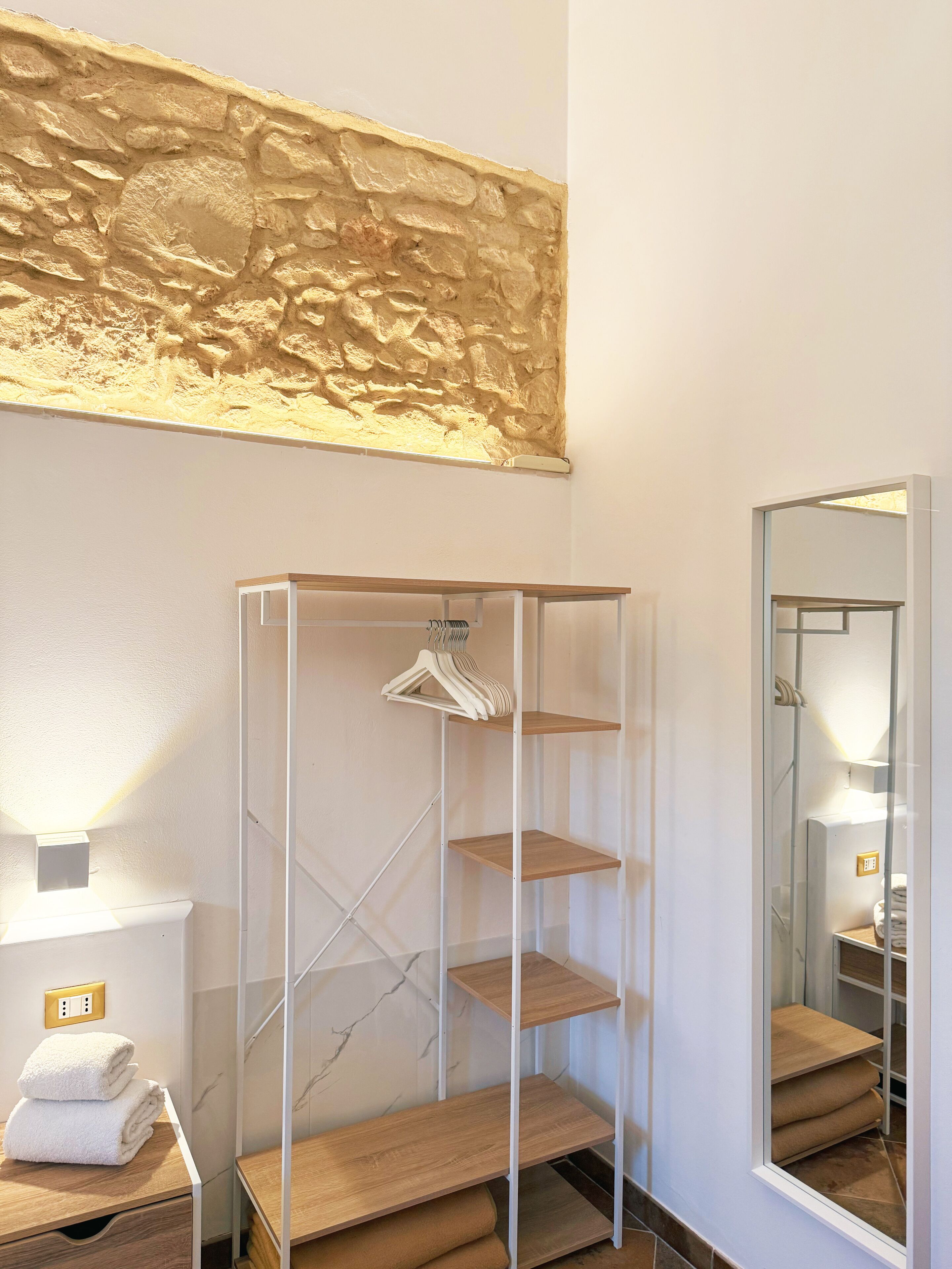 A bathroom with a marble wall, a wooden shelf unit, a mirror, and a towel stack.