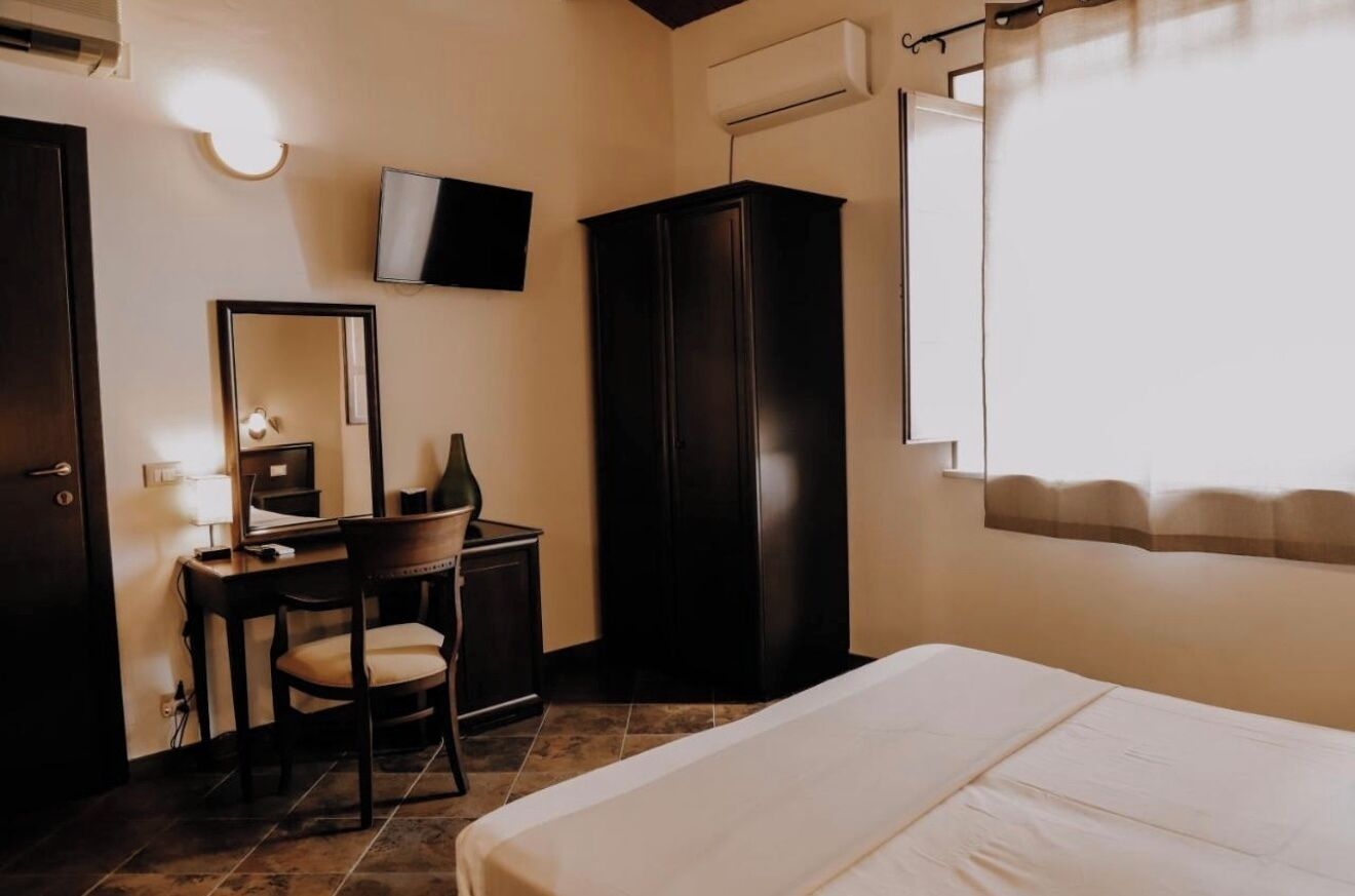 A bedroom with a bed, a desk with a chair, a wardrobe, a TV mounted on the wall, and a window with curtains.