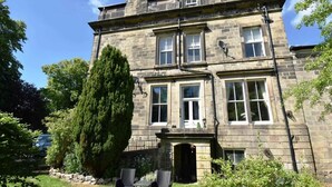 A two-story stone building with a prominent chimney, large windows, and a well-maintained garden with a tall tree and outdoor seating. - Huntress Suite, Buxton - a beautiful victorian apartment with private parking. (Buxton)