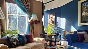 A living room with a beige sofa, a blue sofa, a glass coffee table, and a dining table with chairs. There is a large window with curtains, a floor lamp, and a radiator. - Huntress Suite, Buxton - a beautiful victorian apartment with private parking. (Buxton)