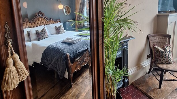 A bedroom with a large mirror, a bed with a dark blue bedspread, a chandelier, and a potted plant. - Huntress Suite, Buxton - a beautiful victorian apartment with private parking. (Buxton)