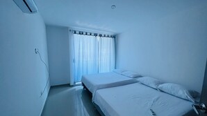 A small, clean room with two single beds, a window with curtains, and a visible electrical outlet on the wall.
