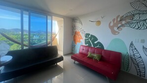 A modern living room with a red sofa and a black couch, large windows with a view of greenery and buildings, and a wall with leaf patterns.