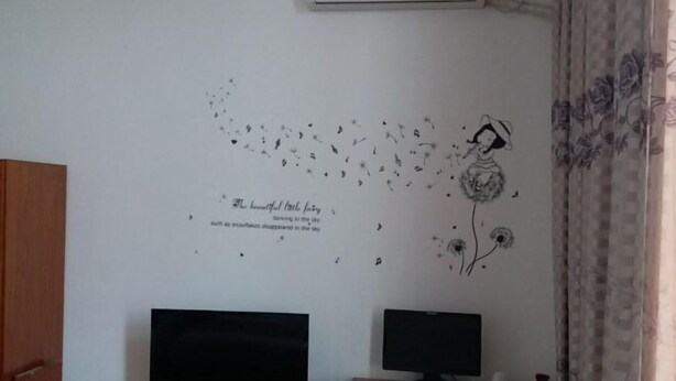 Una habitación con dos monitores en un escritorio, un adhesivo en la pared con una niña y pájaros, y una ventana con cortinas.