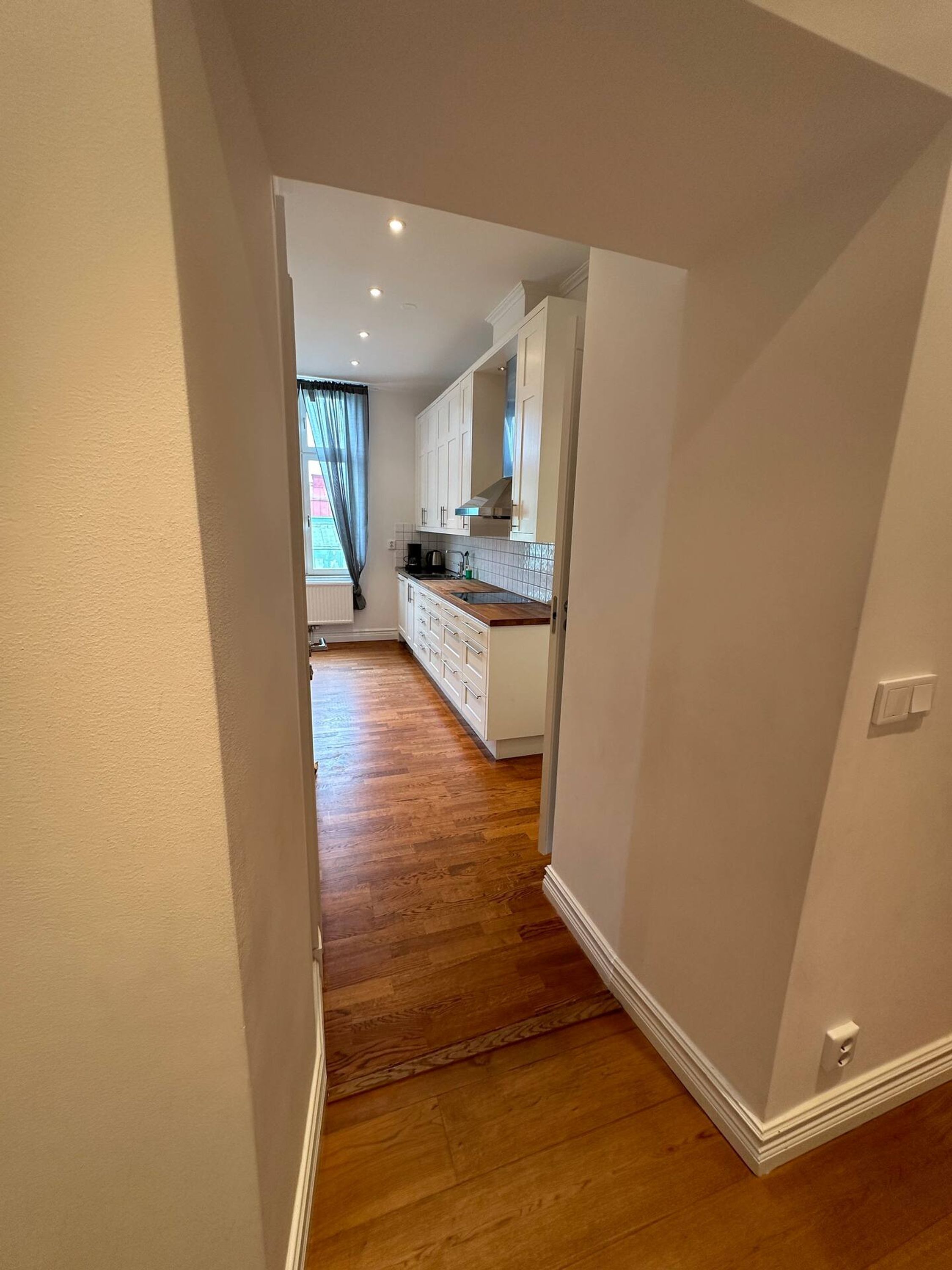 A narrow hallway leading to a kitchen with white cabinets and a wooden floor.