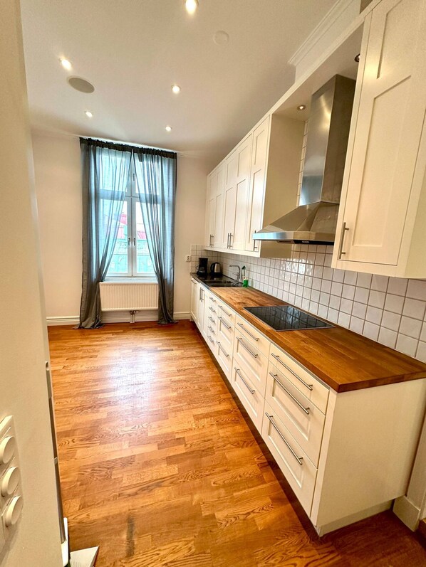 Una cocina con piso de madera, armarios blancos, campana extractora de acero inoxidable y una ventana con cortinas.