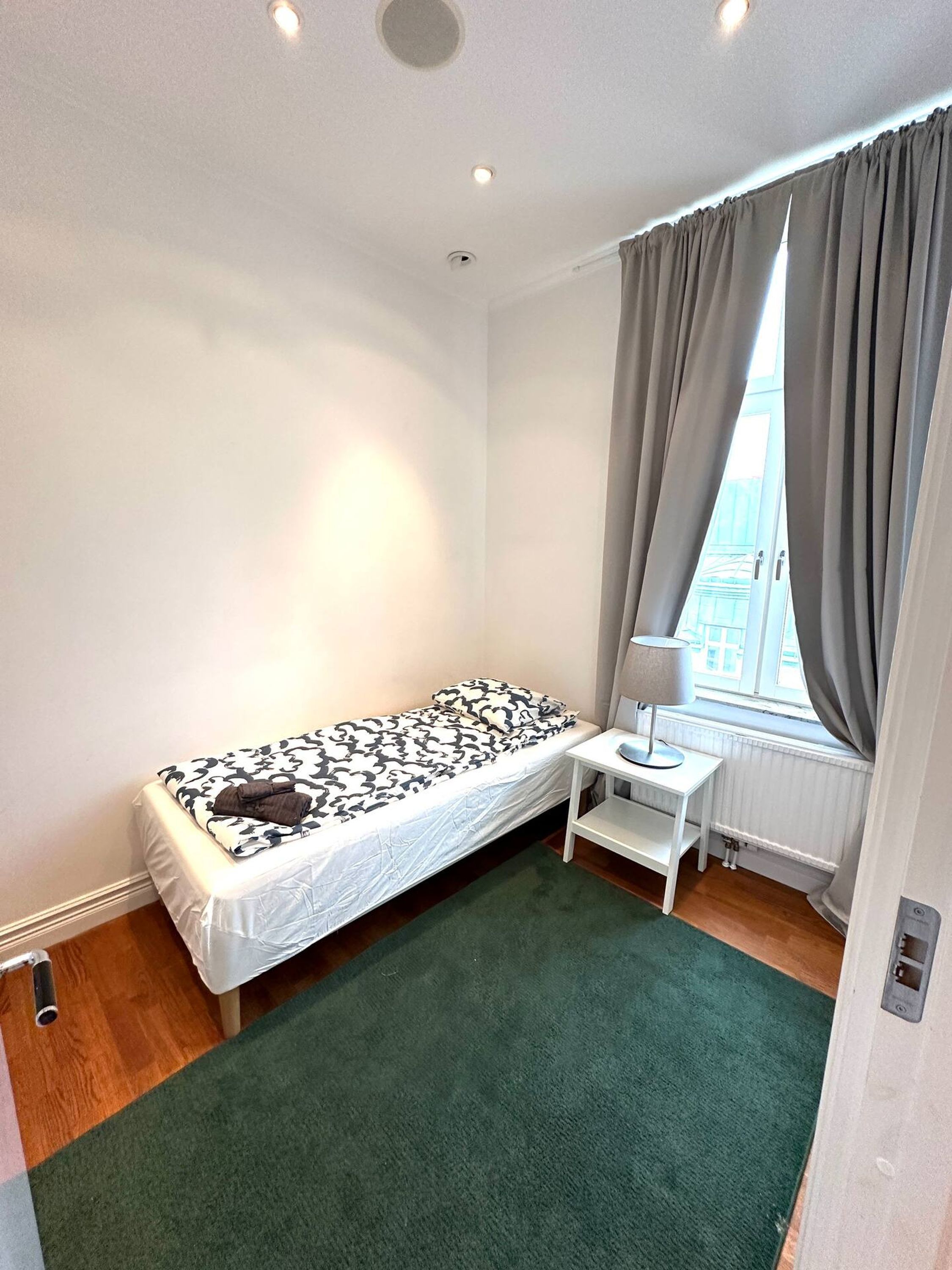 Una habitación pequeña con cama individual, alfombra verde, escritorio blanco con lámpara y ventana con cortinas grises.