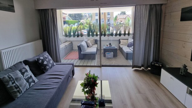 A modern living room with a grey sofa, a coffee table, and a view of a courtyard with plants and a building in the background.
