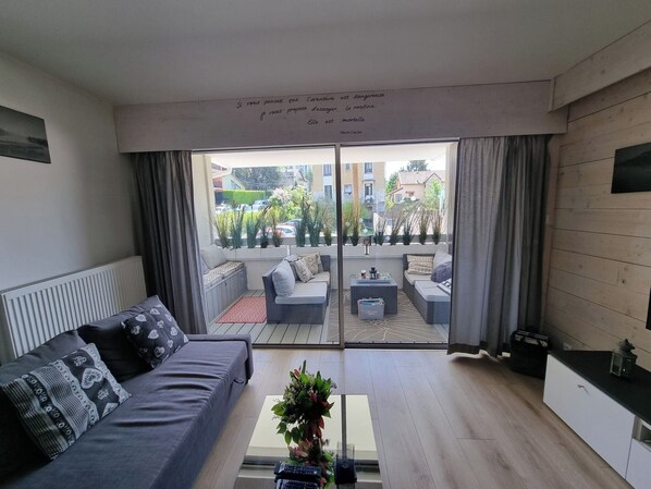 A modern living room with a grey sofa, a coffee table, and a view of a courtyard with plants and a building in the background.