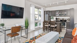 A modern living room with a flat-screen TV, a dining area with wooden chairs, a kitchen with dark cabinets and stainless steel appliances, and a large window with curtains. - Beautiful 1-Bedroom Apartment@Walk to Uptown@Fast WiFi@In-Unit W/D (Minneapolis)